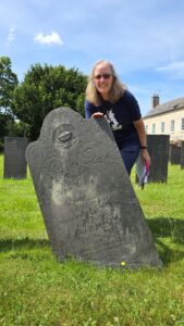 Nancy standing behind Mary Bean Levers’ 1791 headstone in the St. Peter’s church graveyard, with other stones and trees in the background.
